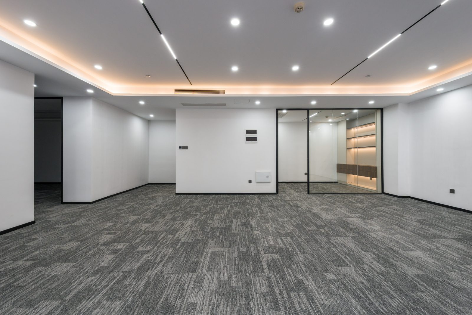Modern empty office space with gray carpet, white walls, recessed ceiling lights, and glass-walled conference room