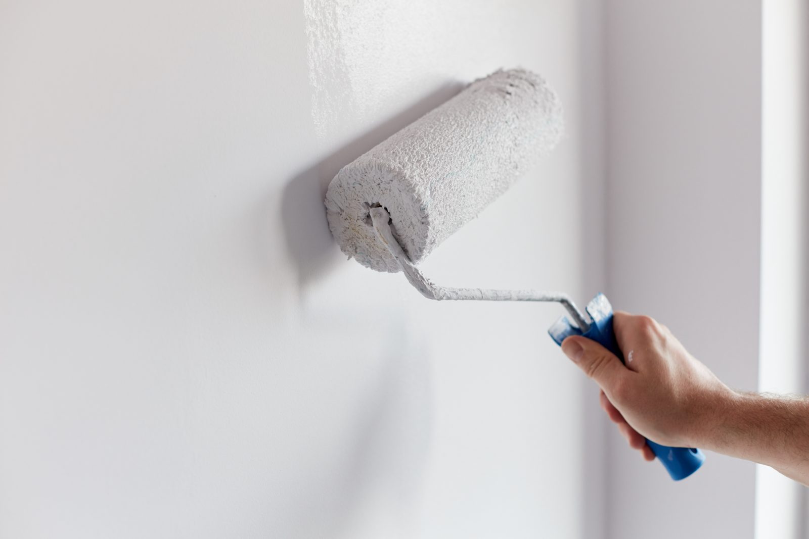 Person painting white wall with paint roller and blue handle